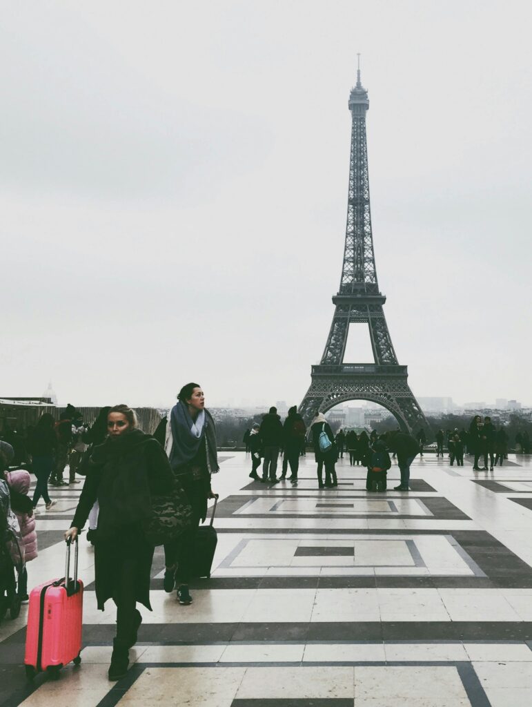 Crowds of tourists with luggage at the Eiffel Tower in Paris on a cloudy day.